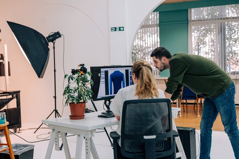 Two colleagues in a photography studio, one seated, one standing, look at an image of a long-sleeved blue top on the computer screen in front of them. 