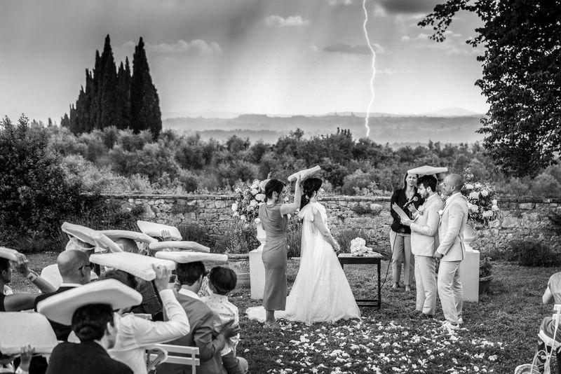 Een bruid en bruidegom geven hun ja-woord. De bruiloftsgasten gebruiken kussens om hun hoofd te bedekken in de regen en in de verte is een bliksemschicht zichtbaar in een zwart-witfoto die is gemaakt door Fabio Mirulla met een Canon EOS R5.