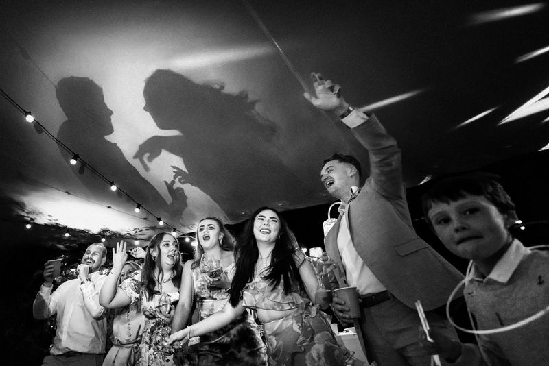 Bruiloftsgasten vieren feest terwijl de schaduw van de bruid en bruidegom op het plafond te zien is, in een zwart-witfoto die is gemaakt door Fabio Mirulla met een Canon EOS R6.