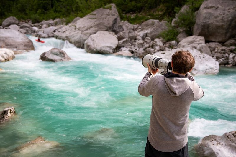   Adventure sports photographer Samo Vidic stands beside rocks above a fast-flowing river photographing a canoeist.