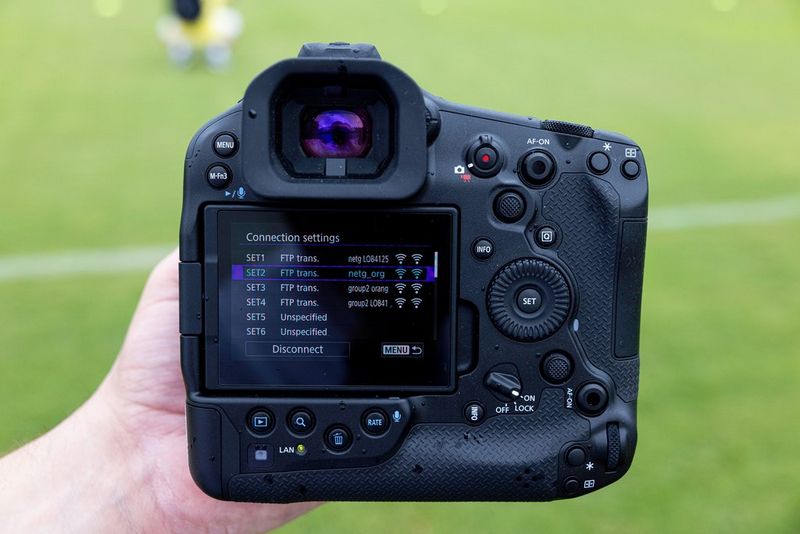 Canon EOS R1 в руках фотографа, стоящего возле поля на стадионе; на камере открыто меню параметров подключения.