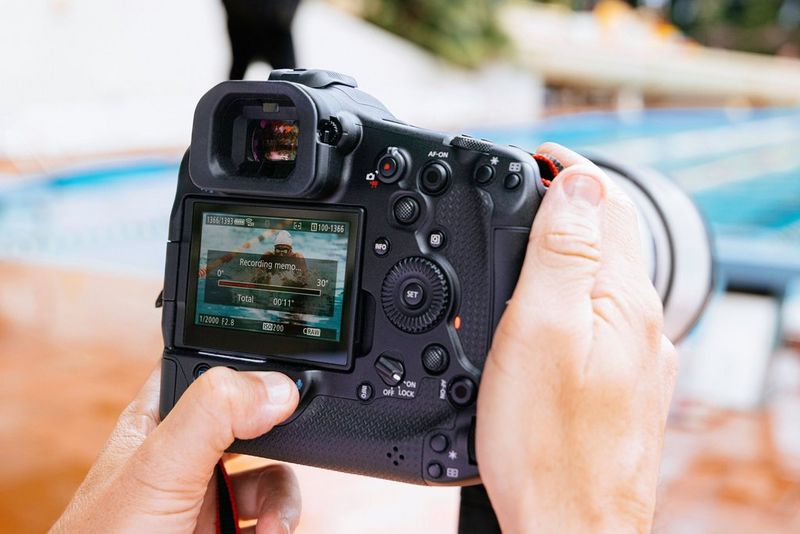 Фотограф спорта возле бассейна записывает голосовую заметку на Canon EOS R1, а на заднем экране камеры можно увидеть фото пловца.
