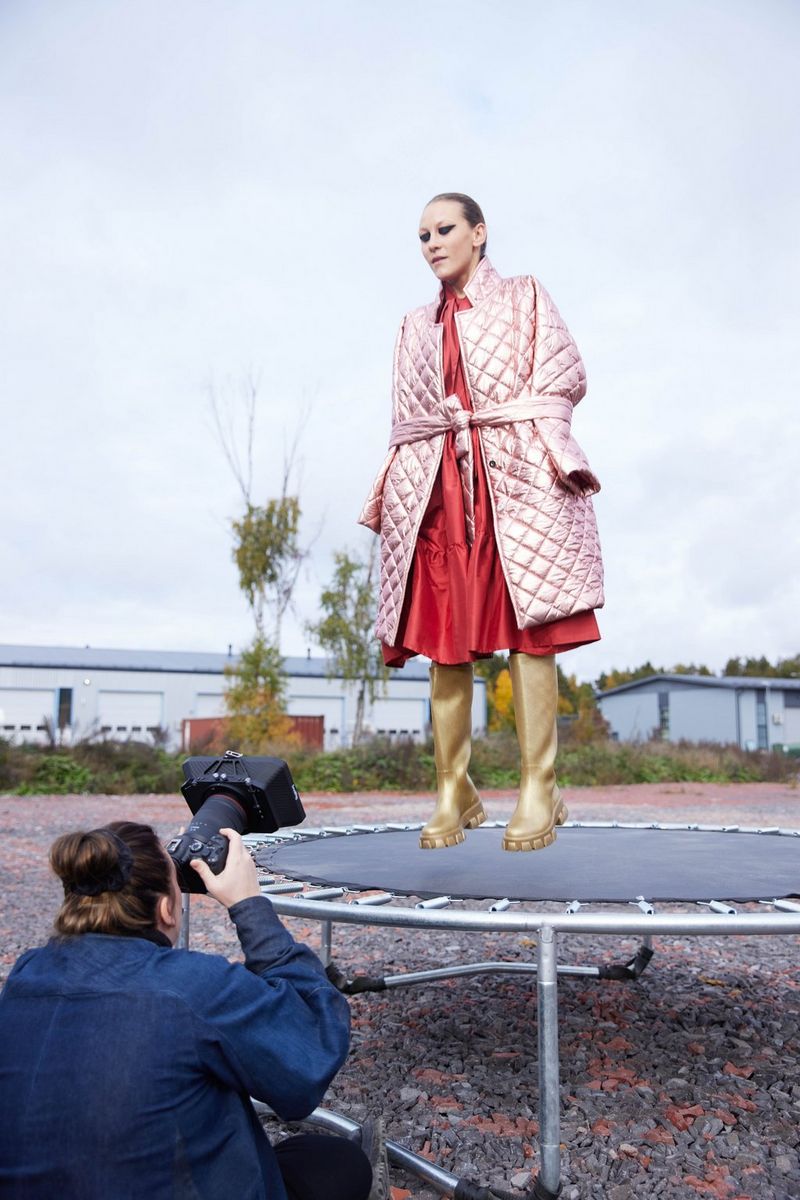 Fotograaf Nana Simelius ligt op de grond met een Canon EOS R6 Mark II in de hand, en maakt opnamen van een model in goudkleurige laarzen, een rode jurk en roze peignoir dat trampolinespringt.