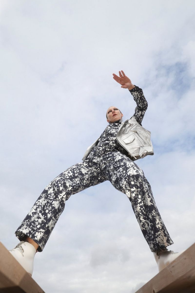 Een model in een jumpsuit met zwart-wit dessin en zilverkleurig gilet, gefotografeerd van beneden af. Het model staat met twee stukken hout tussen haar voeten en houdt één arm omhoog. Gefotografeerd door Nana Simelius met een Canon EOS R6 Mark II.