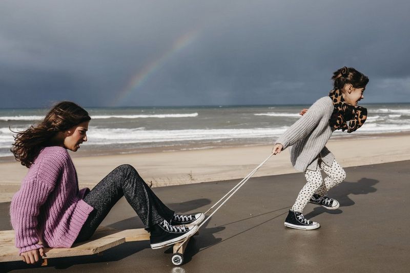 A child is pulled by another child on a board with wheels, the beach and ocean in the background and a rainbow in the sky, in a photo by Marisa Martins taken on a Canon EOS R8.