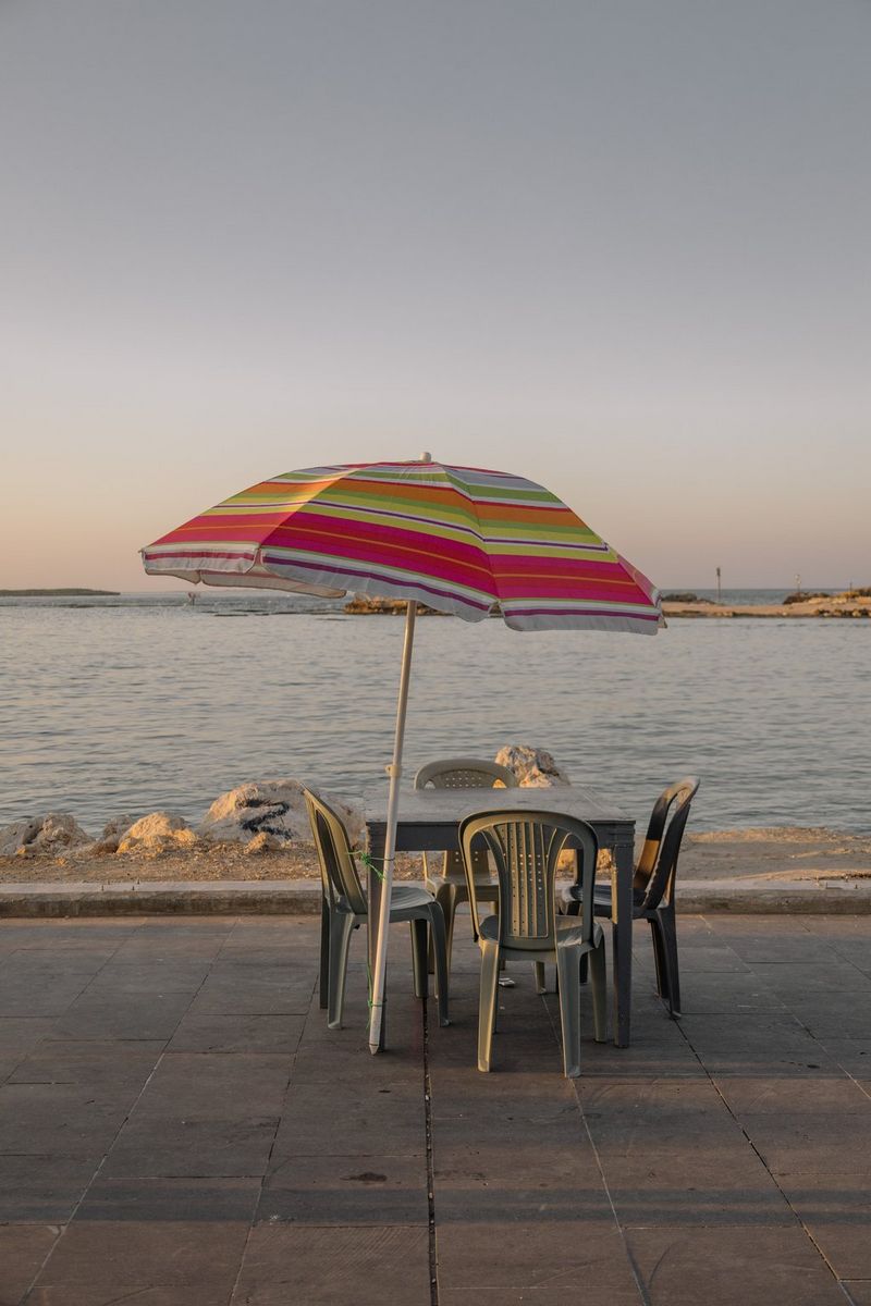 An empty table and chairs with a large umbrella tied to one table leg stand on the seafront in a photo by Aline Deschamps taken on a Canon EOS R8.
