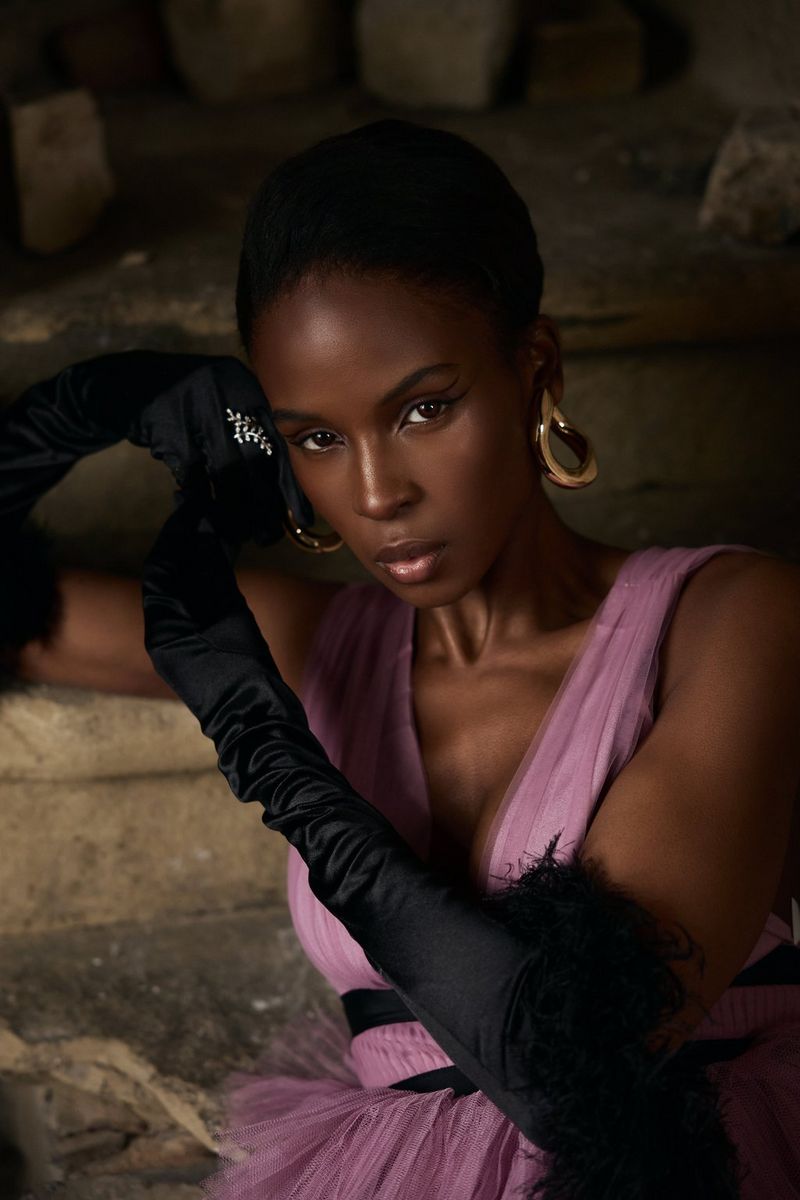 A medium close-up of a person wearing a pink dress and long black gloves leaning against a wall in a photo by Ian Hippolyte, taken on a Canon EOS R8.