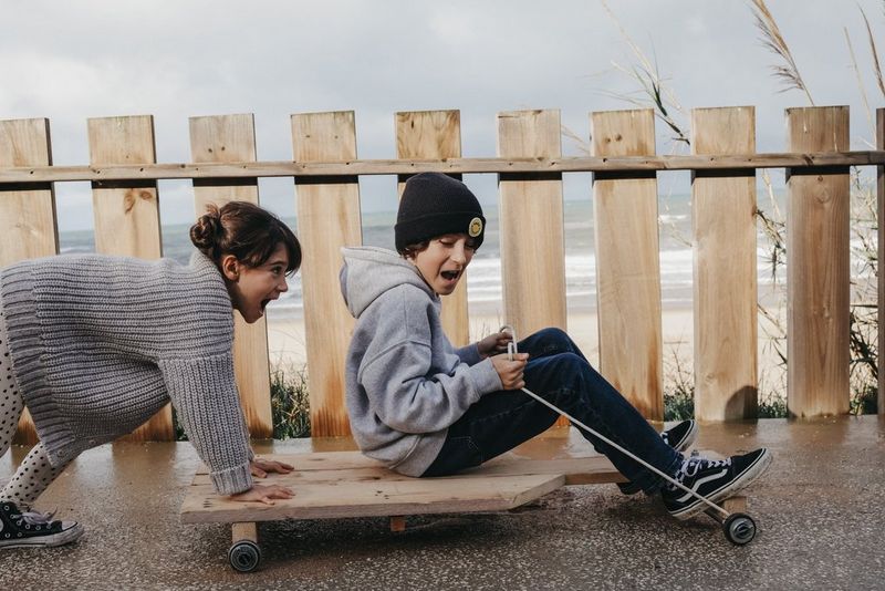 A child pushes another child on a board with wheels next to a fence with the beach in the background, in a photo by Marisa Martins taken on a Canon EOS R8.