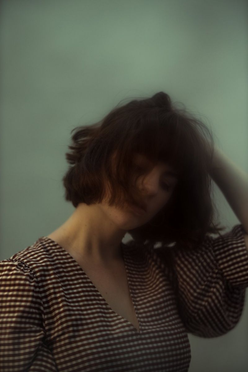 A young woman in a check dress holding her head in anguish, her hair covering much of her face.