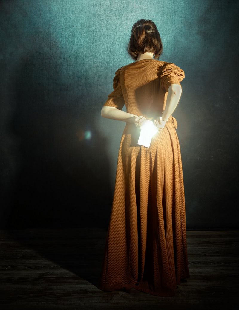A young woman in an orange dress shot from behind, light shining brightly off the phone she holds behind her back.
