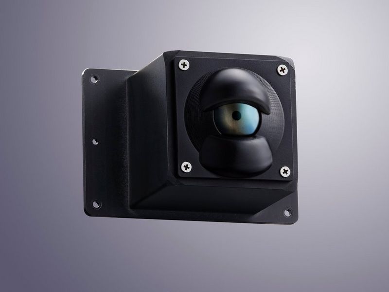 Un piccolo alloggiamento nero con delle viti nei quattro angoli che presenta una bombatura rotonda nella parte anteriore e somiglia a un bulbo oculare con la pupilla al centro.