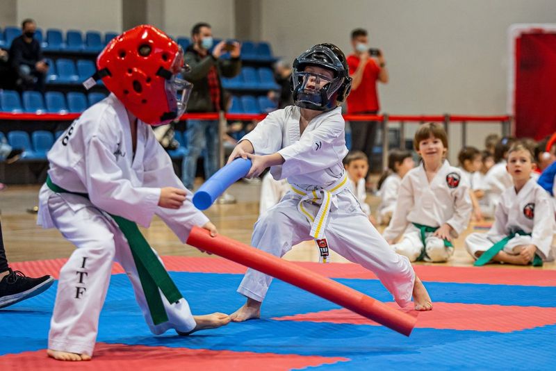 Due giovani atleti di taekwondo che indossano caschi imbottiti combattono con delle aste di gommapiuma, mentre sullo sfondo sfocato sono presenti altri bambini seduti per terra.