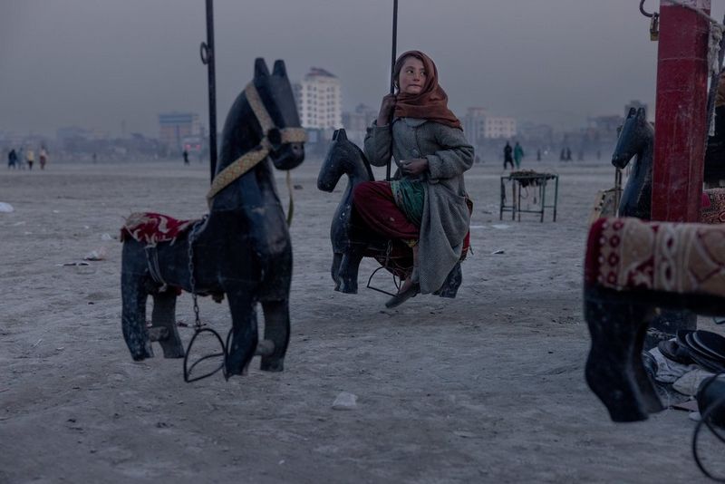 A young girl wearing a scarf over her head and a long grey coat sits on a carousel horse, photographed by Daniel Etter.