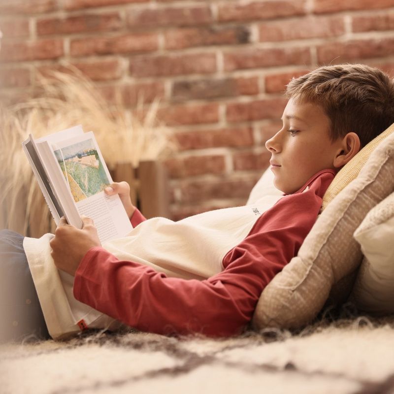 A young boy reads a book while leant against a large stack of cushions.