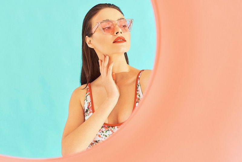 Женщина в розовых солнцезащитных очках стоит перед синей стеной, вид через розовую трубку.