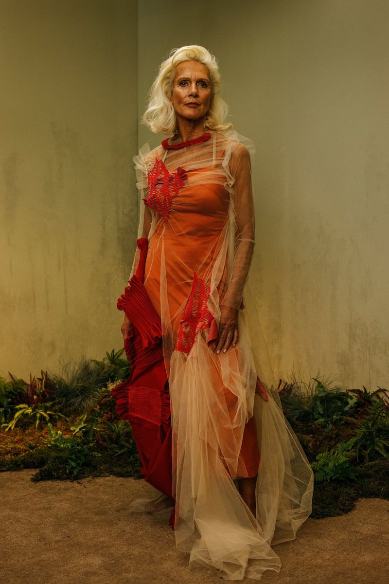 A full-length portrait of an older model wearing an orange dress. 