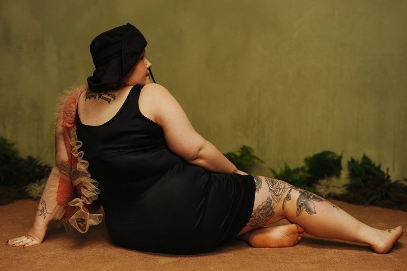 Plus-size, tattooed model Ami is photographed from behind, sitting with her legs to one side in a pose reminiscent of Old Master paintings of reclining models.