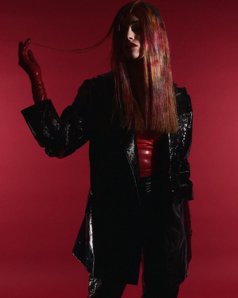 Модель в блестящей черной куртке и длинных красных перчатках позирует на красном фоне, держа в одной руке несколько прядей своих длинных каштановых волос.