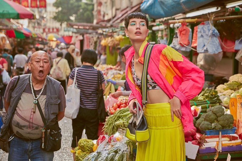 A fashion model in a bright yellow skirt and a fluorescent pink jacket poses at a busy market, while an open-mouthed man walks alongside her.