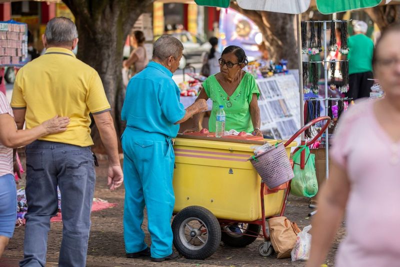 A man in a blue shirt and trousers and a woman in a green top stand talking over a food cart in a busy market square.