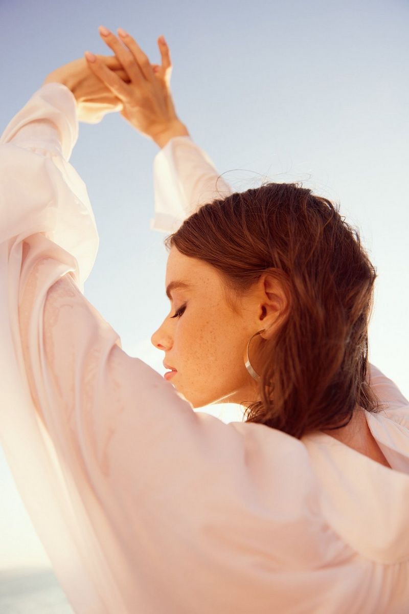 A woman wearing a floaty white top stands outside, raising her arms above her head and facing away from the camera with her head turned slightly towards it and her eyes closed. Taken by Jaroslav Monchak on a Canon EOS R5.