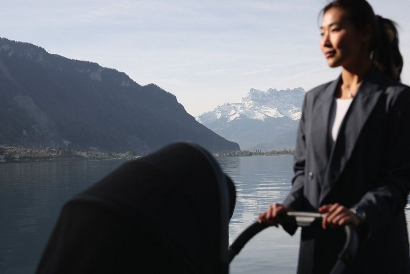 A woman wearing a suit pushing a pram is at the front of the frame, out of focus, while behind her and in focus are a lake and two mountains. Taken by Jaroslav Monchak on a Canon EOS R5.