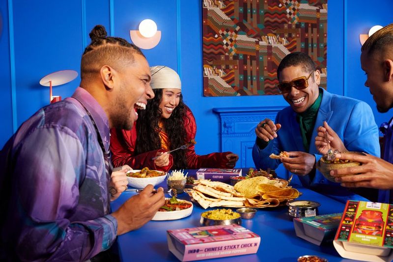 Sur cette photo prise par Kid Circus, quatre personnes habillées de couleurs vives mangent autour d'une table, sur un fond bleu éclatant.