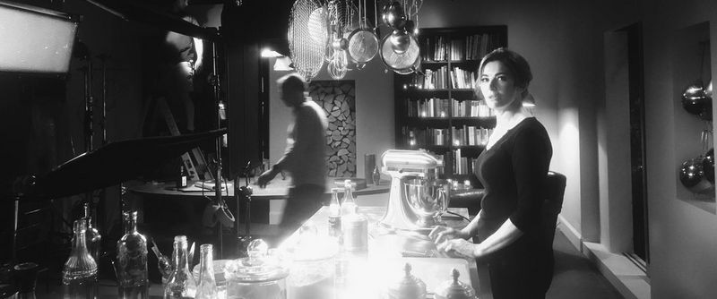 Nigella Lawson è in piedi davanti al bancone della cucina rivolta verso la videocamera, mentre i membri della troupe sistemano delle luci sullo sfondo.