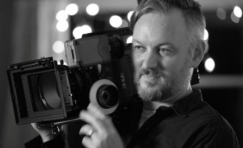 Il direttore della fotografia Robin Fox con una videocamera poggiata sulla spalla destra.