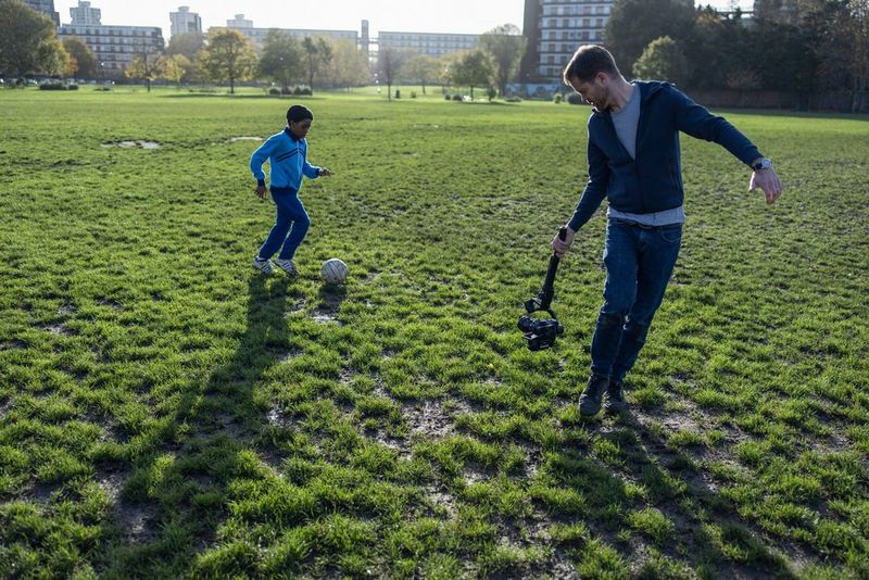 Оператор-постановщик Патрик Смит держит в руке закрепленную на ручном подвесе-стабилизаторе кинокамеру Canon EOS C70 и снимает ребенка, который играет в футбол.