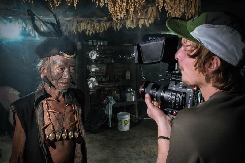 Filmmaker Michael Zomer films an indigenous man with tattoos covering his face and chest in a small hut with a Canon camera. 