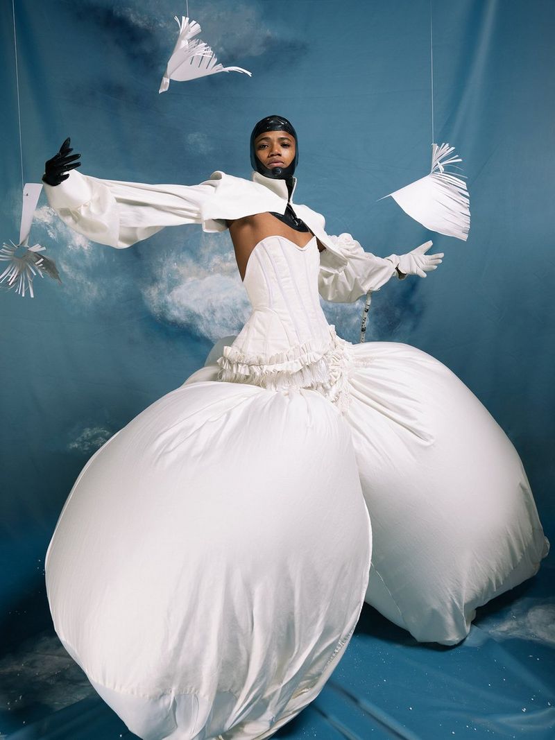 Una modella che indossa un abito bianco con una gonna gonfiabile e un passamontagna in PVC nero con le braccia aperte su uno sfondo blu.
