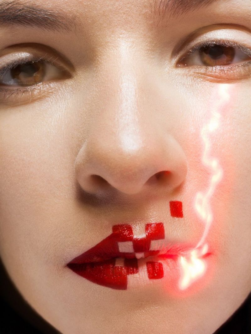 Un primo piano del viso di una donna; ha un disegno geometrico rosso sulle labbra e una scia di luce che va dalla bocca all'occhio.