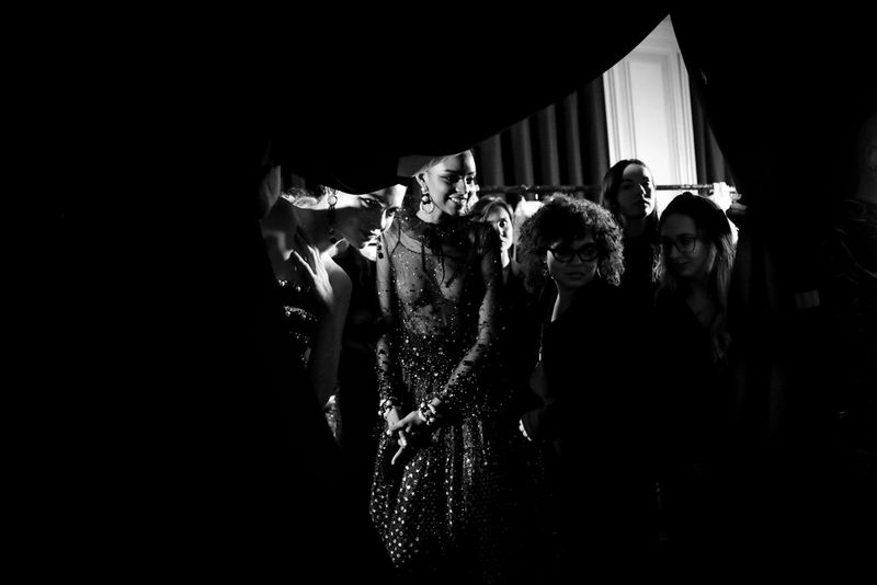 A black-and-white shot of models watching a fashion show from behind a curtain in their dressing room.