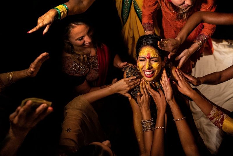 Женщина в южноазиатском наряде стоит среди толпы женщин, которые тянутся к ней и наносят на ее лицо желтую краску.