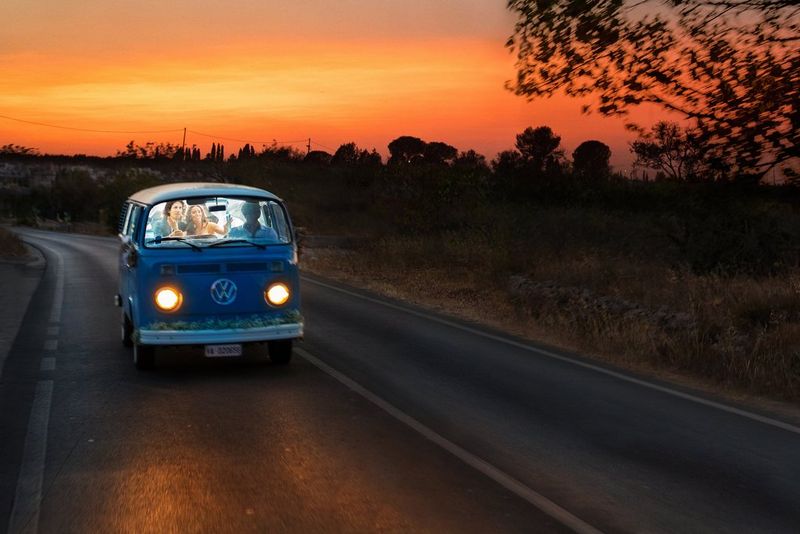 Молодожены на заднем диване дома на колесах от VW едут по проселочной дороге на фоне заката.