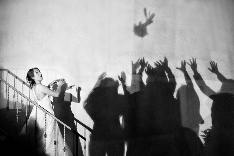 Черно-белое изображение, на котором невеста удивленно смотрит вверх, стоя на лестнице. Тени на стене возле нее позволяют представить группу женщин, которые пытаются поймать брошенный букет.