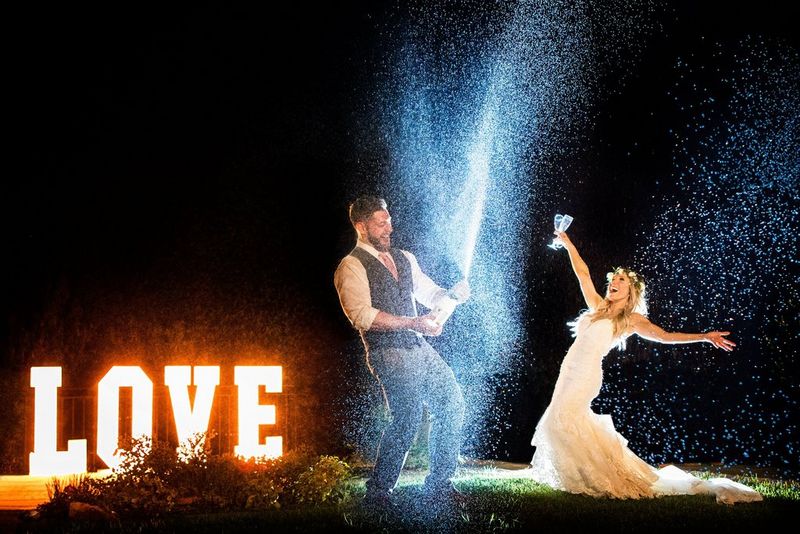 Мужчина открывает шампанское, повернувшись в сторону невесты, — подсвеченные вспышкой брызги летят в воздухе, а рядом ярким светом горит неоновая вывеска LOVE.