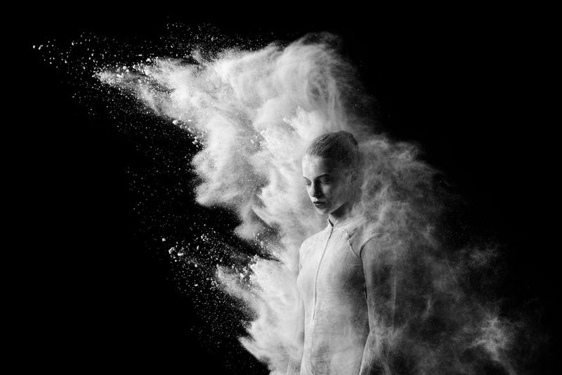 A black and white image of a ballet dancer standing stationary while a cloud of flour billows above her from behind. 