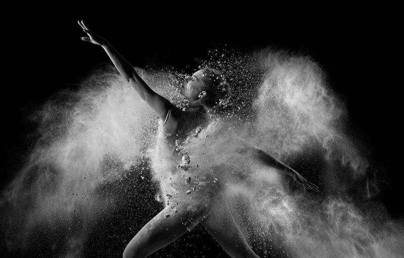 A dancer moving through a cloud of flour, one arm stretched out in front and one behind, with large particles visible in the foreground.