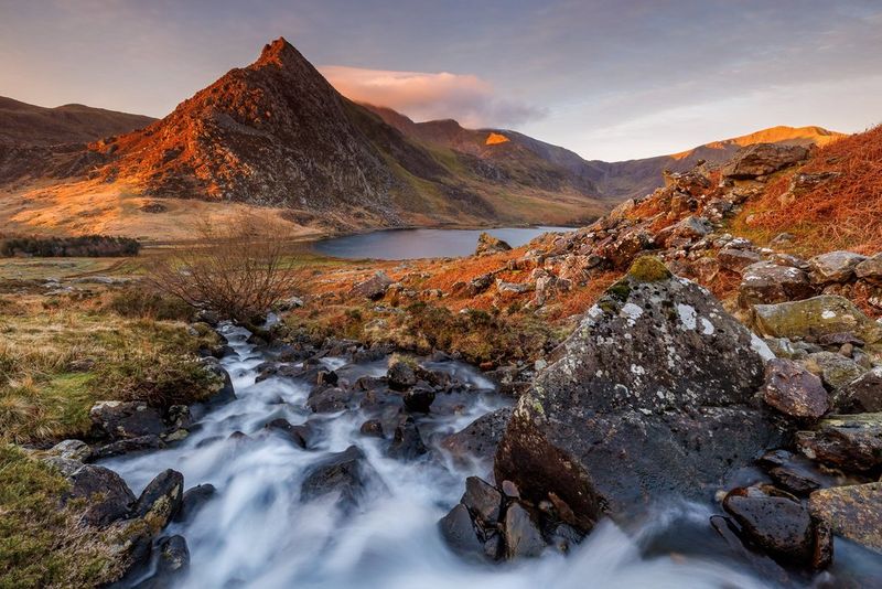 A focus-stacked image of running water flowing towards a lake surrounded by vegetation of a rich orange hue in a photograph by Chris Ceaser.