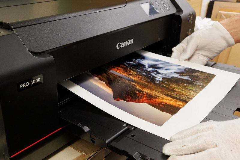 A pair of hands, wearing white gloves, removes a landscape print from a Canon imagePROGRAF PRO-1000 printer.