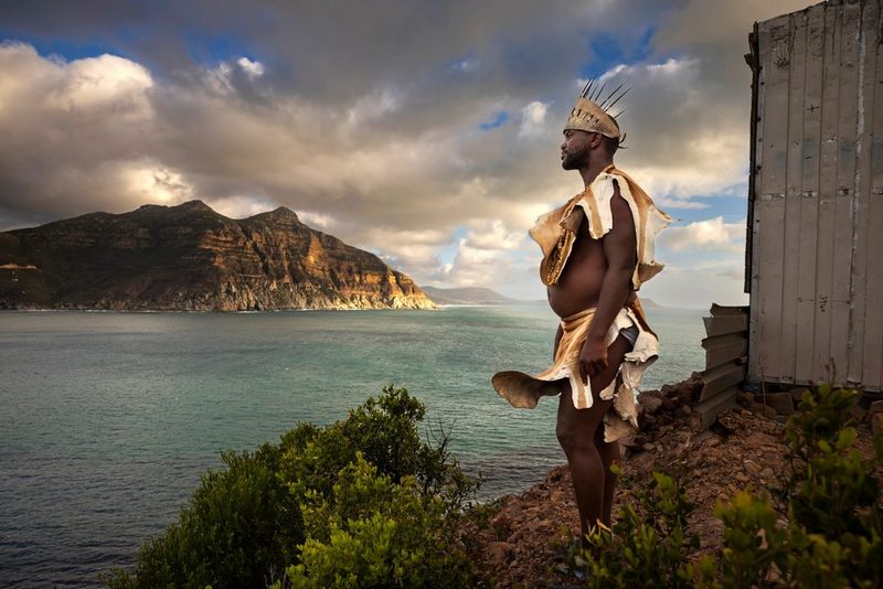 Un uomo in abiti tribali guarda il mare. Alla sua destra c'è una piccola capanna e sullo sfondo una vasta catena montuosa. Scatto realizzato da Brent Stirton con Canon EOS R5.