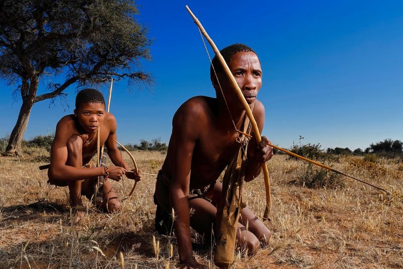 Due giovani che indossano abiti tribali e portano archi e frecce fatti a mano accovacciati tra l'erba ingiallita. Scatto realizzato da Brent Stirton con Canon EOS R.