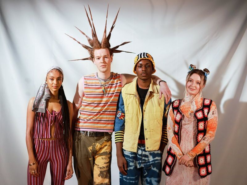 A group shot of four young people in bold outfits, the tallest man has hair spiked up in different directions.