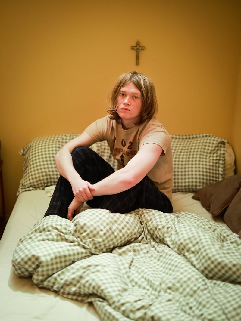 A young man with hair to his shoulders stares at the camera while sitting on a bed. Behind him is a mustard yellow wall with a small wooden cross on it.