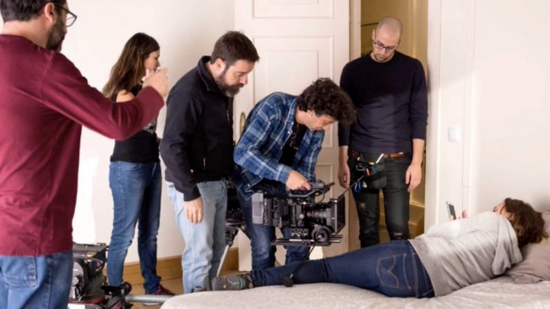 Juan Luis Cabellos directs a cinematographer filming a woman lying on a bed looking at her mobile phone, with other crew members on set.