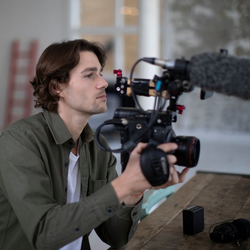 A person looks into a camera, holding it aloft to the right of the shot.