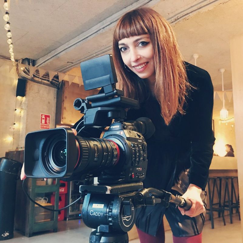 Cinematographer Irene Cruz leans forward, smiling, holding the handle of the tripod of her Canon cine camera.