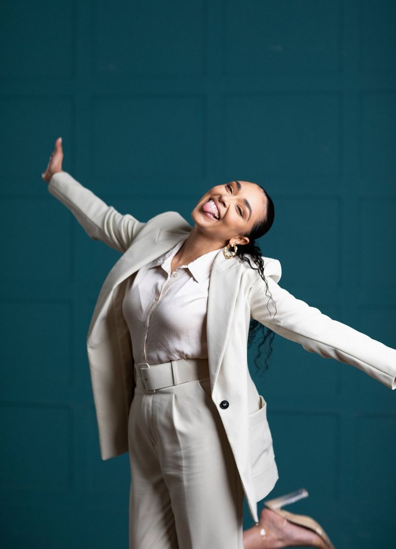 Ashleigh smiles and sticks out her tongue as she stands on one leg with her arms outstretched, wearing a smart white and cream suit.